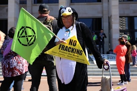 A person dressed in a penguin costume holds a green flag with a black symbol and a yellow triangular sign that reads 'NO FRACKING. Protect Our Water. Clean Country - Healthy Community.' The person appears to wear glasses and a headpiece with braids. There are other people in the background, some dressed casually and others in costumes. The scene seems to take place in front of a building with steps.