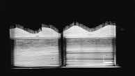 Two windows with blinds partially closed are illuminated from behind, creating sharp contrasts of light and shadow. The blinds are slightly open, casting horizontal lines across the scene. Above, the wavy edges of a lace curtain add a soft texture to the otherwise sharp geometric lines.