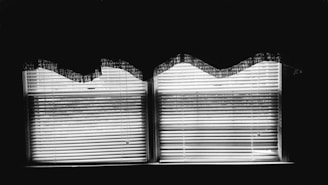 Two windows with blinds partially closed are illuminated from behind, creating sharp contrasts of light and shadow. The blinds are slightly open, casting horizontal lines across the scene. Above, the wavy edges of a lace curtain add a soft texture to the otherwise sharp geometric lines.