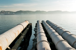 Several large pipelines run parallel to each other over a body of water with hilly terrain in the background. The pipelines show signs of rust and weathering. The water is calm and reflects the subtle morning or evening light, creating a serene atmosphere.