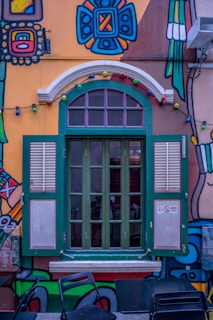 A vibrant, colorful mural decorates a building wall with a large, green-framed window featuring shutters. The artwork includes geometric and abstract patterns in bold colors, along with a string of multicolored lights hanging above the window. There are black chairs and a table positioned in front of the window, and a small 'No Smoking' sign is visible.