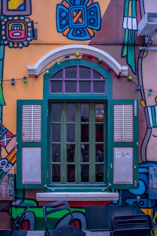 A vibrant, colorful mural decorates a building wall with a large, green-framed window featuring shutters. The artwork includes geometric and abstract patterns in bold colors, along with a string of multicolored lights hanging above the window. There are black chairs and a table positioned in front of the window, and a small 'No Smoking' sign is visible.
