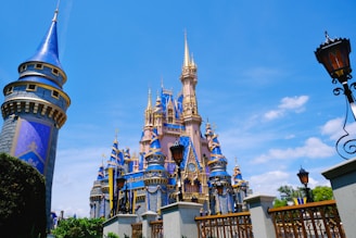 A grand and colorful fairy-tale castle with tall golden spires and royal blue rooftops, surrounded by decorative details. The sky is bright blue with a few clouds, adding to the picturesque scene. Tall evergreen trees and ornamental street lamps frame the view.