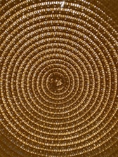 A close-up view of a woven pattern forming concentric circles, with a texture that resembles braided or knitted material in a golden hue. The repetitive circular design creates a mesmerizing and visually symmetrical effect.