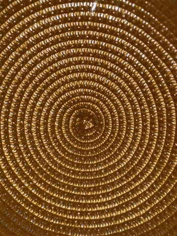 A close-up view of a woven pattern forming concentric circles, with a texture that resembles braided or knitted material in a golden hue. The repetitive circular design creates a mesmerizing and visually symmetrical effect.