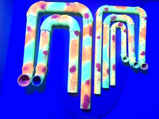 A collection of pipes against a blue background, painted with bright colors including neon green, orange, and pink. The pipes are arranged in a visually interesting pattern and appear to be glowing under a blacklight.