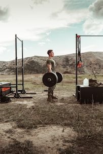 Man lifting weights outdoors with a focused expression, surrounded by nature.
