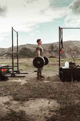 A determined person lifting weights outdoors surrounded by tall pine trees under a clear blue sky.