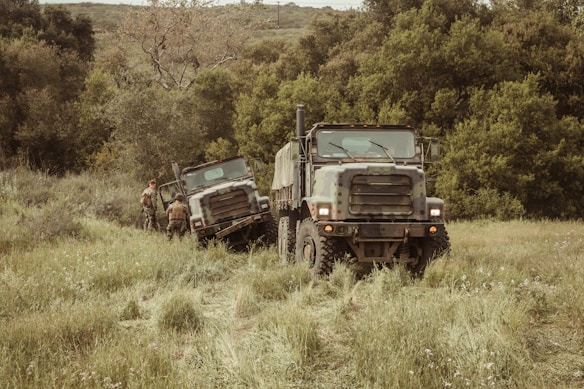 Two military-style trucks are parked on a grassy field surrounded by dense green foliage. Two individuals in camouflage apparel stand near the trucks engaged in activity. The scene suggests a rural or remote setting with natural, untouched vegetation.