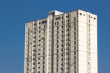 A tall, rectangular apartment building with multiple floors, featuring beige and white exterior walls set against a bright blue sky. The building has small windows evenly distributed across each floor, and several air conditioning units are visible.