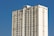 A tall, rectangular apartment building with multiple floors, featuring beige and white exterior walls set against a bright blue sky. The building has small windows evenly distributed across each floor, and several air conditioning units are visible.