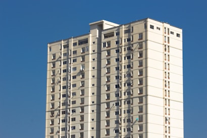 A tall, rectangular apartment building with multiple floors, featuring beige and white exterior walls set against a bright blue sky. The building has small windows evenly distributed across each floor, and several air conditioning units are visible.