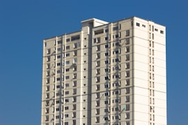 A tall, rectangular apartment building with multiple floors, featuring beige and white exterior walls set against a bright blue sky. The building has small windows evenly distributed across each floor, and several air conditioning units are visible.