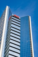 A tall modern skyscraper with many windows reflecting the blue sky. The building has a sleek, glass facade and is labeled with the red letters 'AON' near the top. The architecture is sharp and geometric, creating a sense of height and modernity.