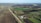 Aerial view of expansive farmland, featuring various plots with dark soil and green crops. A dirt road runs through the fields, with rows of tall trees lining parts of the road. Small farm buildings are visible near the trees. The landscape stretches into the distance with an open sky.