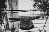 A black and white image capturing a puffer jacket hanging on metal railing bars, with a perspective looking upwards. The background includes parts of a building with visible lines of electrical wires and tree foliage on one side.