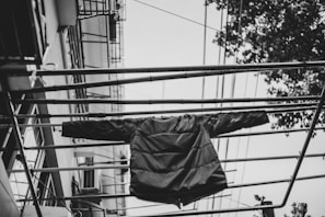 A black and white image capturing a puffer jacket hanging on metal railing bars, with a perspective looking upwards. The background includes parts of a building with visible lines of electrical wires and tree foliage on one side.
