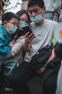 A group of three people wearing face masks are closely gathered around a smartphone, appearing to focus intently on the screen. They are outdoors, and the adults are seated. The young child is in a blue jacket, leaning in to look at the device. The man is wearing a gray t-shirt with a printed design and appears to be holding a black jacket or bag.