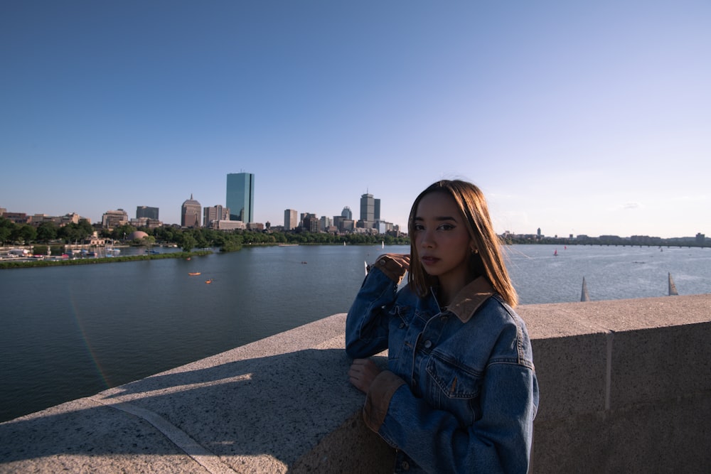 woman in blue denim jacket sitting on gray concrete pavement near body of water during daytime