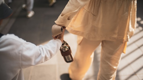 A close-up view captures a person in a beige outfit holding hands with a child dressed in white. The focus is on their clasped hands, conveying a sense of connection and warmth. Soft lighting casts gentle shadows on the ground, giving a warm and intimate feel to the scene.