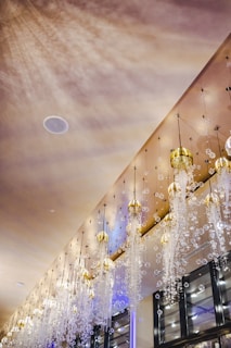 An elegant interior ceiling adorned with a series of intricate chandeliers hanging down. These chandeliers have golden caps and are embellished with numerous sparkling crystal-like beads cascading downwards. The ambient lighting creates a warm glow, casting soft shadows on the ceiling, while the reflections add to the luxurious ambiance.