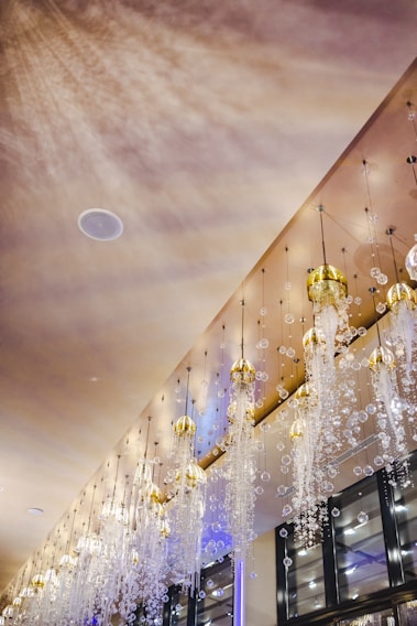 An elegant interior ceiling adorned with a series of intricate chandeliers hanging down. These chandeliers have golden caps and are embellished with numerous sparkling crystal-like beads cascading downwards. The ambient lighting creates a warm glow, casting soft shadows on the ceiling, while the reflections add to the luxurious ambiance.