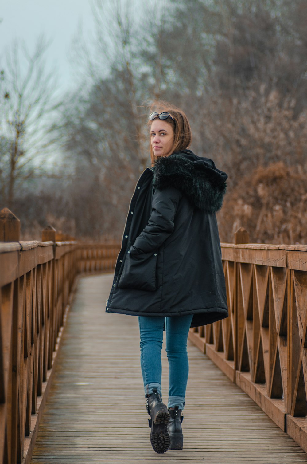 woman in black coat and blue denim jeans standing on wooden bridge during daytime