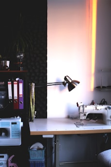 A well-organized sewing workspace with a sewing machine on a wooden table. The area is illuminated by a desk lamp with folders, measuring tapes, and other sewing supplies neatly arranged. The wall has soundproofing foam, adding to the focused and serene atmosphere.