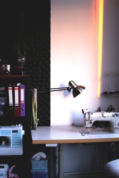 A well-organized sewing workspace with a sewing machine on a wooden table. The area is illuminated by a desk lamp with folders, measuring tapes, and other sewing supplies neatly arranged. The wall has soundproofing foam, adding to the focused and serene atmosphere.