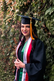 A young woman smiles while wearing a graduation cap and gown. She is adorned with multiple colorful stoles and cords, and stands next to a wall covered in lush green ivy.
