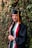 A young woman smiles while wearing a graduation cap and gown. She is adorned with multiple colorful stoles and cords, and stands next to a wall covered in lush green ivy.