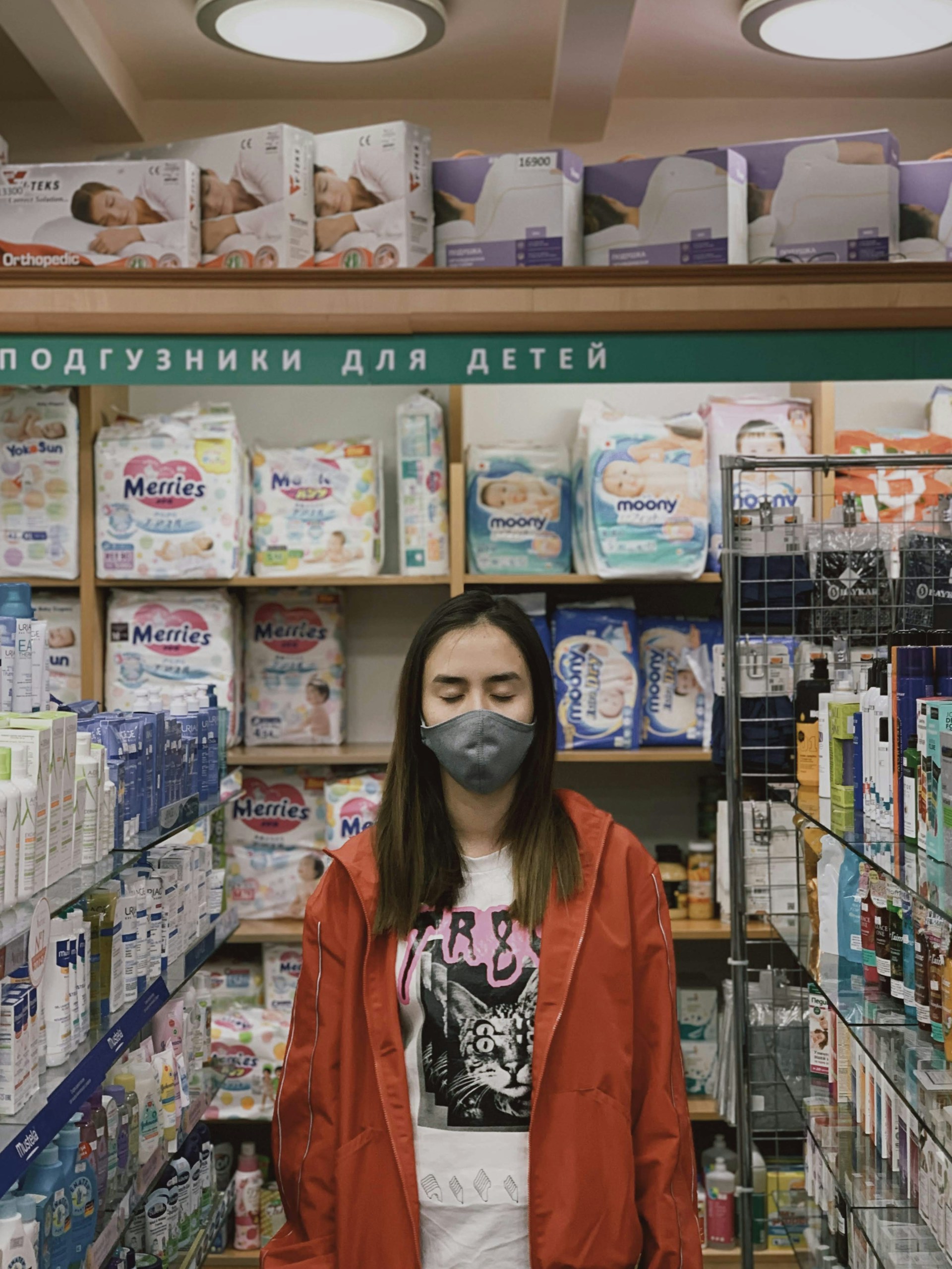 A person is standing in a store aisle wearing a red jacket and a face mask. They have their eyes closed. The shelves are stocked with various baby products like diapers and skincare items. The section is labeled in Cyrillic script, which suggests it is a baby care aisle.
