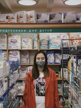 A person is standing in a store aisle wearing a red jacket and a face mask. They have their eyes closed. The shelves are stocked with various baby products like diapers and skincare items. The section is labeled in Cyrillic script, which suggests it is a baby care aisle.
