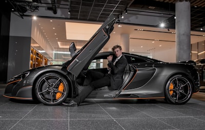 A luxury sports car with its driver-side door open, revealing a man in a suit seated inside. The sleek vehicle features a streamlined design with orange accents on the wheels and body. The setting appears to be a modern showroom with overhead lighting and reflective surfaces.