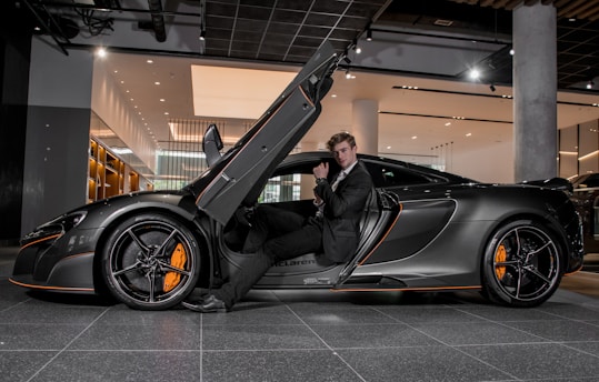 A luxury sports car with its driver-side door open, revealing a man in a suit seated inside. The sleek vehicle features a streamlined design with orange accents on the wheels and body. The setting appears to be a modern showroom with overhead lighting and reflective surfaces.