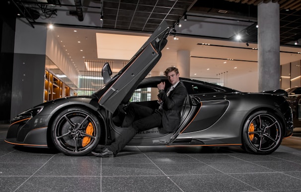 A luxury sports car with its driver-side door open, revealing a man in a suit seated inside. The sleek vehicle features a streamlined design with orange accents on the wheels and body. The setting appears to be a modern showroom with overhead lighting and reflective surfaces.