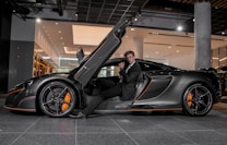 A luxury sports car with its driver-side door open, revealing a man in a suit seated inside. The sleek vehicle features a streamlined design with orange accents on the wheels and body. The setting appears to be a modern showroom with overhead lighting and reflective surfaces.