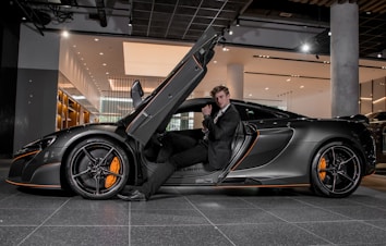 A luxury sports car with its driver-side door open, revealing a man in a suit seated inside. The sleek vehicle features a streamlined design with orange accents on the wheels and body. The setting appears to be a modern showroom with overhead lighting and reflective surfaces.