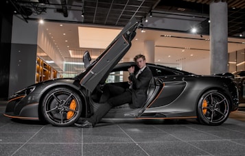 A luxury sports car with its driver-side door open, revealing a man in a suit seated inside. The sleek vehicle features a streamlined design with orange accents on the wheels and body. The setting appears to be a modern showroom with overhead lighting and reflective surfaces.