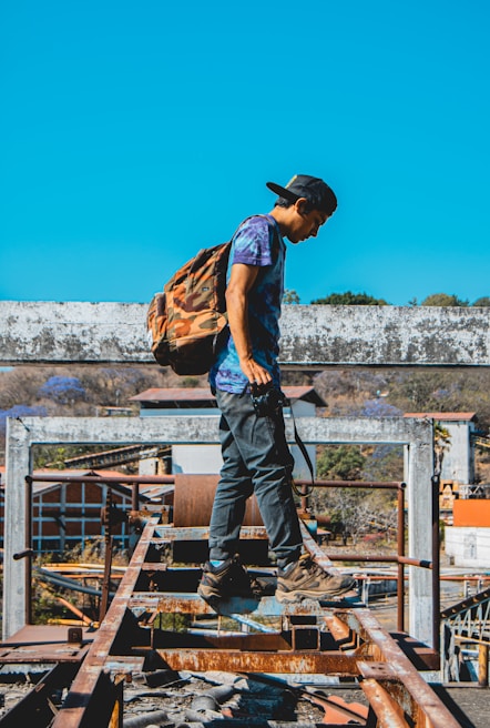 A person wearing a camouflage backpack, cap, and casual clothing is standing on a rusty metal structure outdoors, with a bright blue sky in the background. They appear to be exploring or adventuring in an industrial or construction-like setting. Some buildings and trees are visible in the distance.