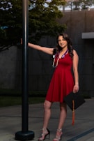 A woman in a red dress stands outdoors next to a black lamppost. She is holding a graduation cap with an orange tassel in one hand and is wearing a stethoscope around her neck. Her other arm rests on the lamppost. The background features a concrete wall and greenery.