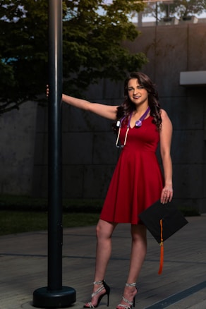 A woman in a red dress stands outdoors next to a black lamppost. She is holding a graduation cap with an orange tassel in one hand and is wearing a stethoscope around her neck. Her other arm rests on the lamppost. The background features a concrete wall and greenery.