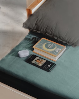 A neatly arranged bedside setup includes a pillow, a stack of two books with a decorative cover, a pair of wireless earbuds in their case, and a smartphone displaying a music player interface. The setting appears calm and organized with soft light casting shadows across the fabric surface.