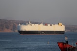 A large cargo ship is sailing on a calm body of water, with a mountainous landscape in the background under a clear sky. The ship is predominantly white with a dark hull and features various equipment on its deck. In the foreground, part of another smaller red vessel is visible, along with a flag.