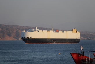 A large cargo ship is sailing on a calm body of water, with a mountainous landscape in the background under a clear sky. The ship is predominantly white with a dark hull and features various equipment on its deck. In the foreground, part of another smaller red vessel is visible, along with a flag.