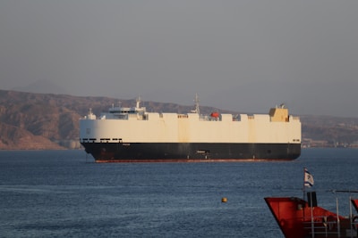 A large cargo ship is sailing on a calm body of water, with a mountainous landscape in the background under a clear sky. The ship is predominantly white with a dark hull and features various equipment on its deck. In the foreground, part of another smaller red vessel is visible, along with a flag.