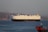 A large cargo ship is sailing on a calm body of water, with a mountainous landscape in the background under a clear sky. The ship is predominantly white with a dark hull and features various equipment on its deck. In the foreground, part of another smaller red vessel is visible, along with a flag.