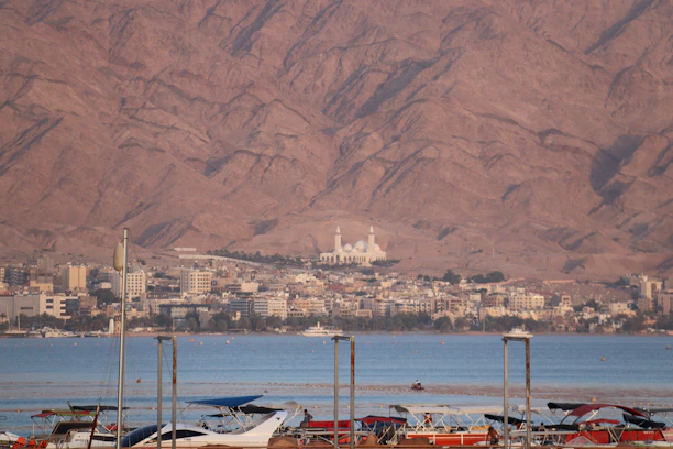 A coastal cityscape with a marina in the foreground, featuring boats docked on calm blue waters. Behind the marina, a city with diverse architecture extends across the landscape, with a prominent mosque featuring multiple domes visible near the center. The backdrop consists of impressive, rugged mountains with a reddish hue under natural sunlight.