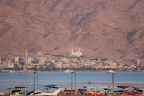 A coastal cityscape with a marina in the foreground, featuring boats docked on calm blue waters. Behind the marina, a city with diverse architecture extends across the landscape, with a prominent mosque featuring multiple domes visible near the center. The backdrop consists of impressive, rugged mountains with a reddish hue under natural sunlight.