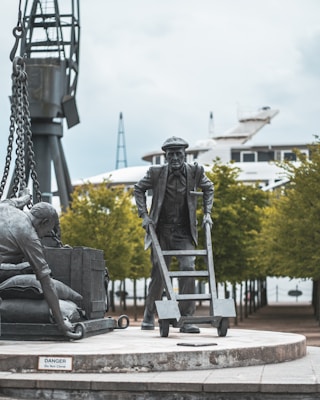 A sculpture of two figures engaged in work, one using a dolly and the other lifting a crate. The setting is outdoors with trees lining a pathway in the background. A large industrial crane and a ship are visible behind the figures, suggesting a maritime or dockside location.
